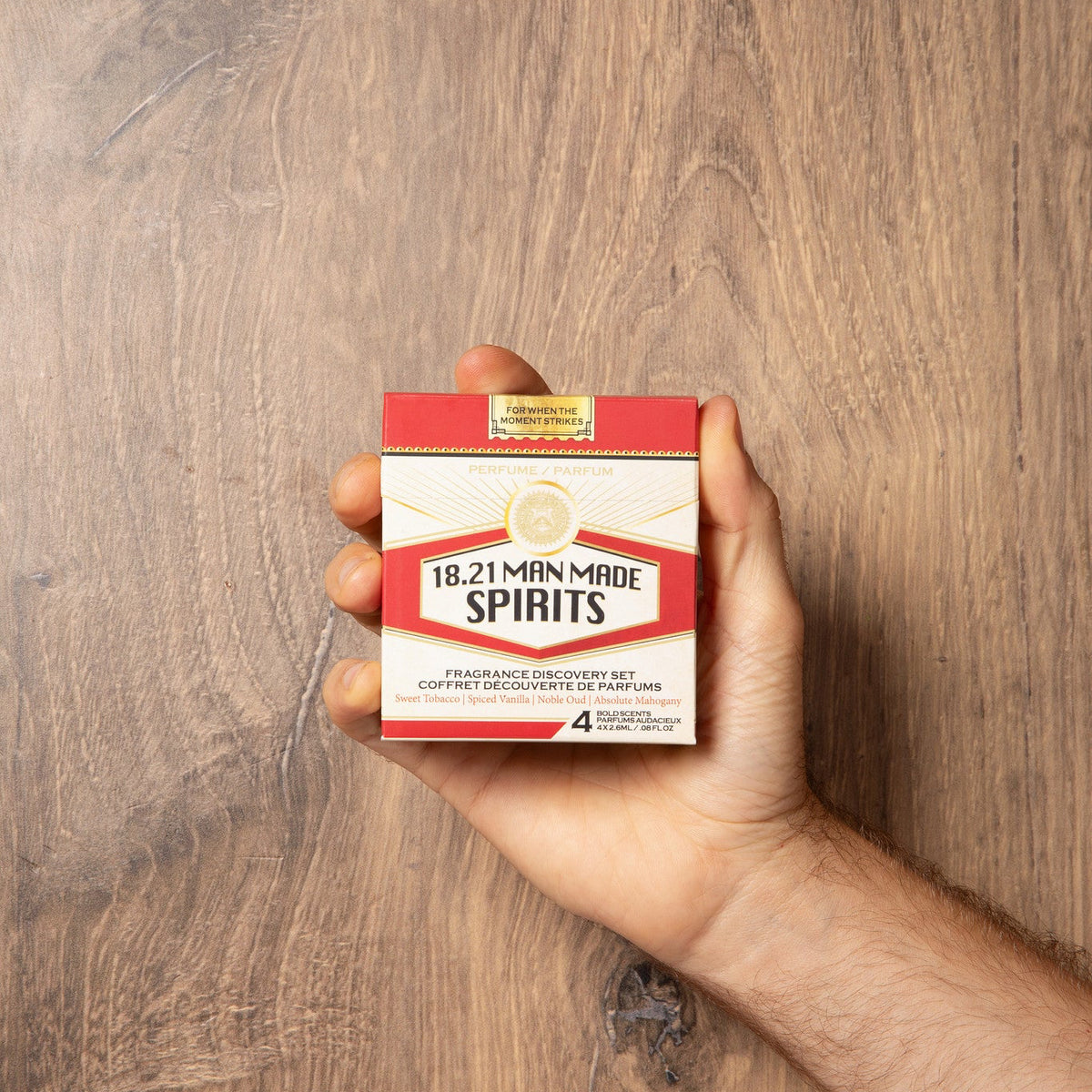 A hand holding a small box of 18.21 Minute Spirits cologne discover set against a brown wood-grain background.