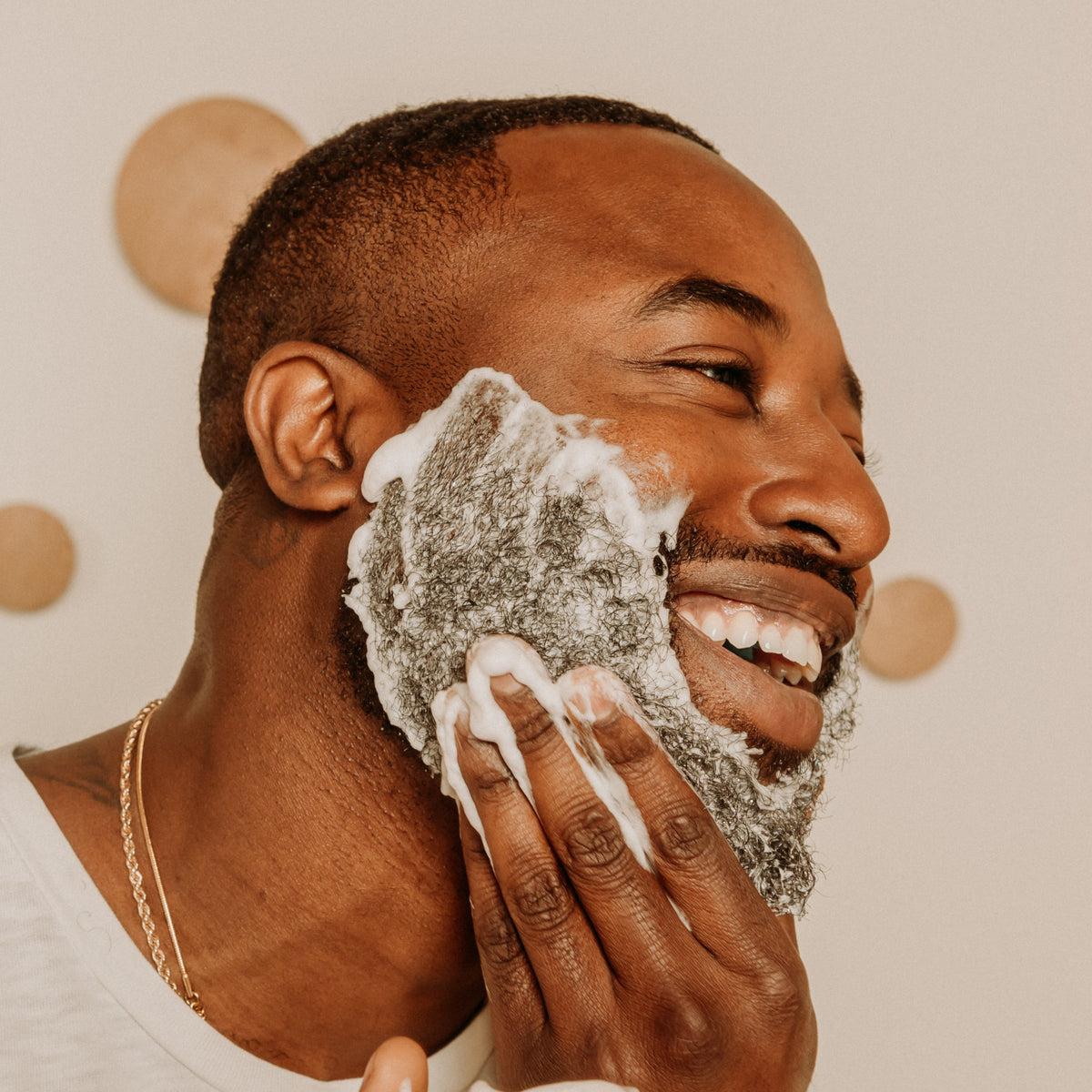 A close-up lifestyle shot of a man washing his face with face and beard wash in sweet tobacco wearing a white t-shirt and a gold chain necklace against a light beige background with circular wall decorations.