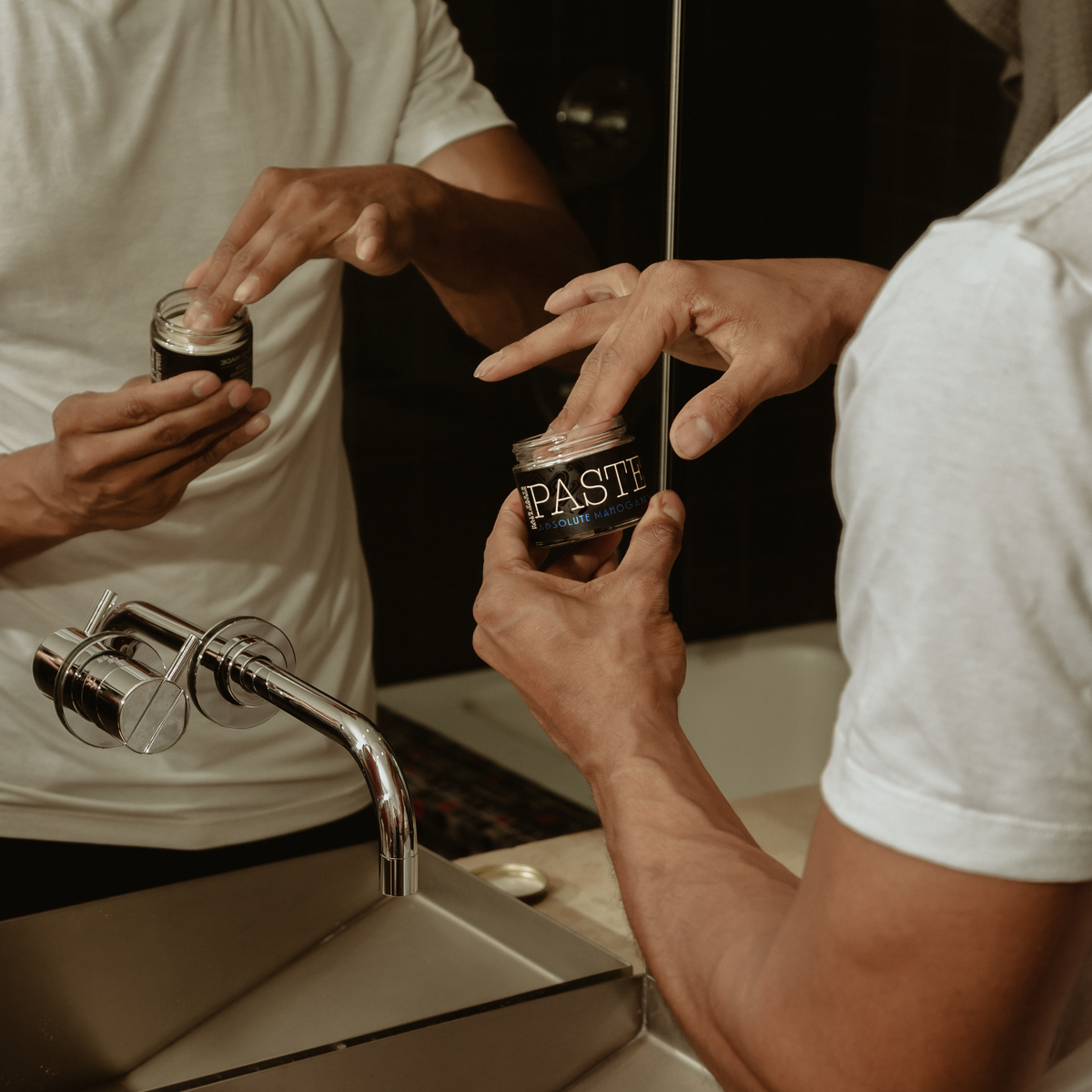 Person applying hair product from a jar labeled 'PASTE' in front of a mirror.