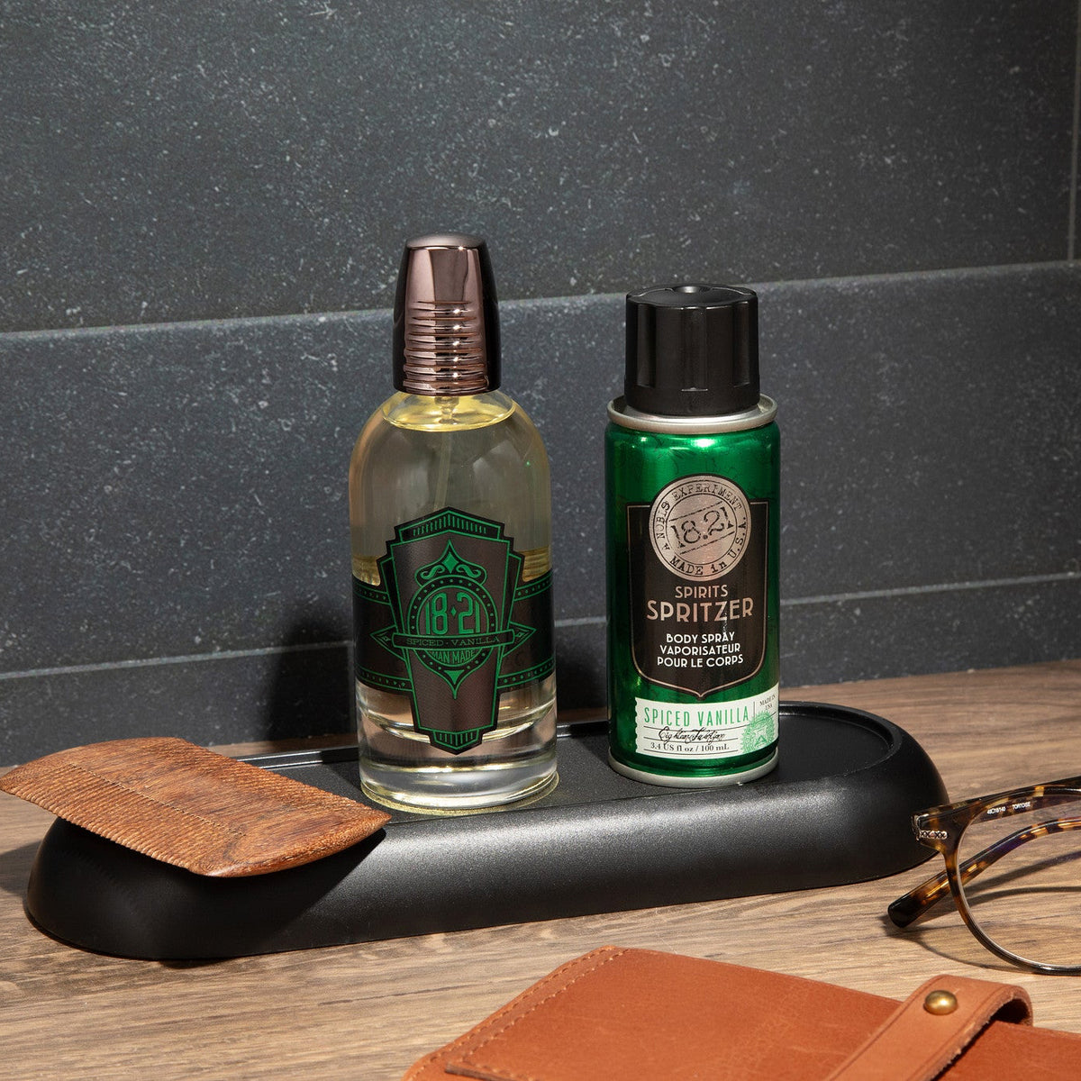 Two bottles, cologne and spritzer on a wooden surface with a dark tiled wall background.