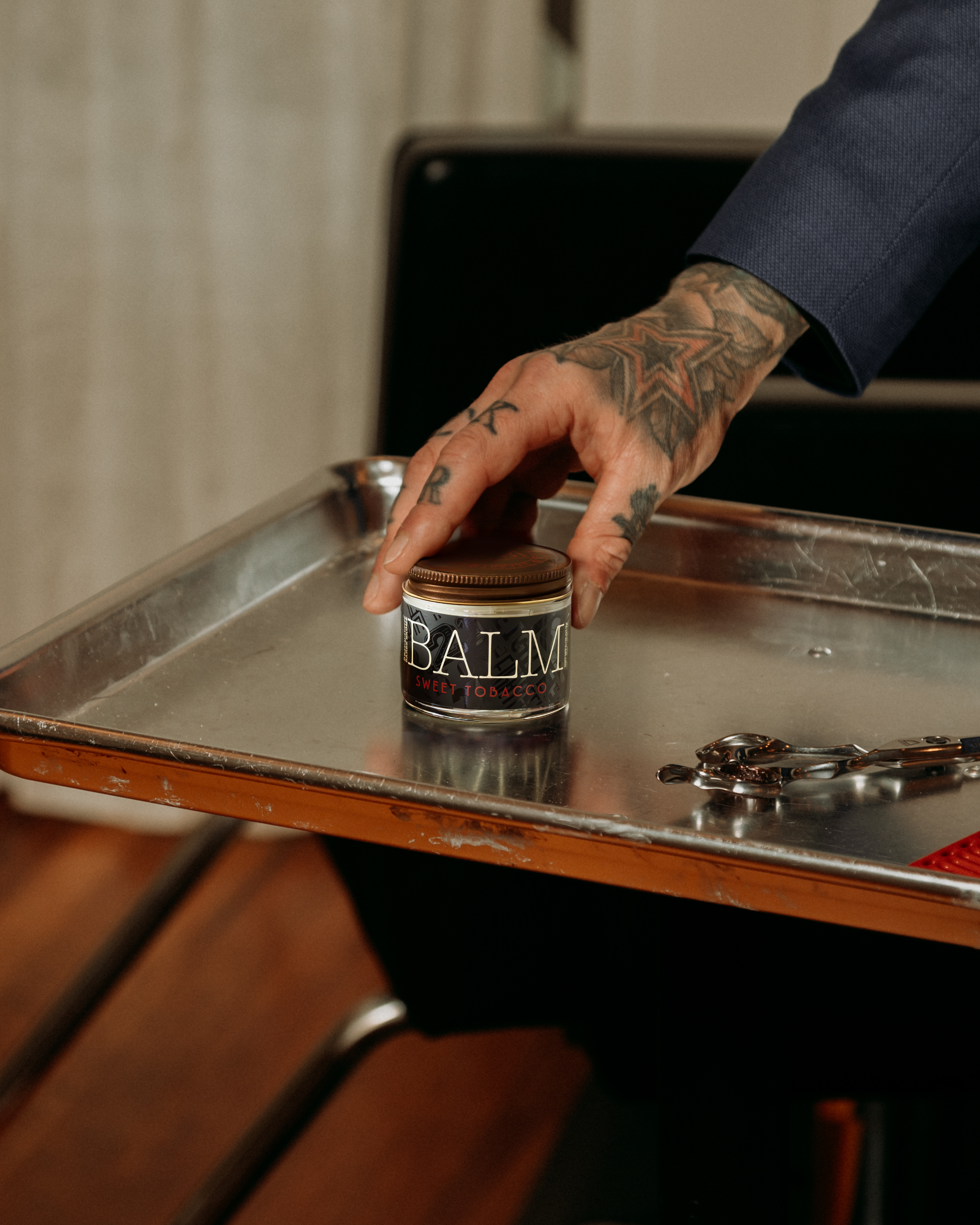 Person with tattooed hand reaching for a jar labeled 'Balm' on a metallic surface.