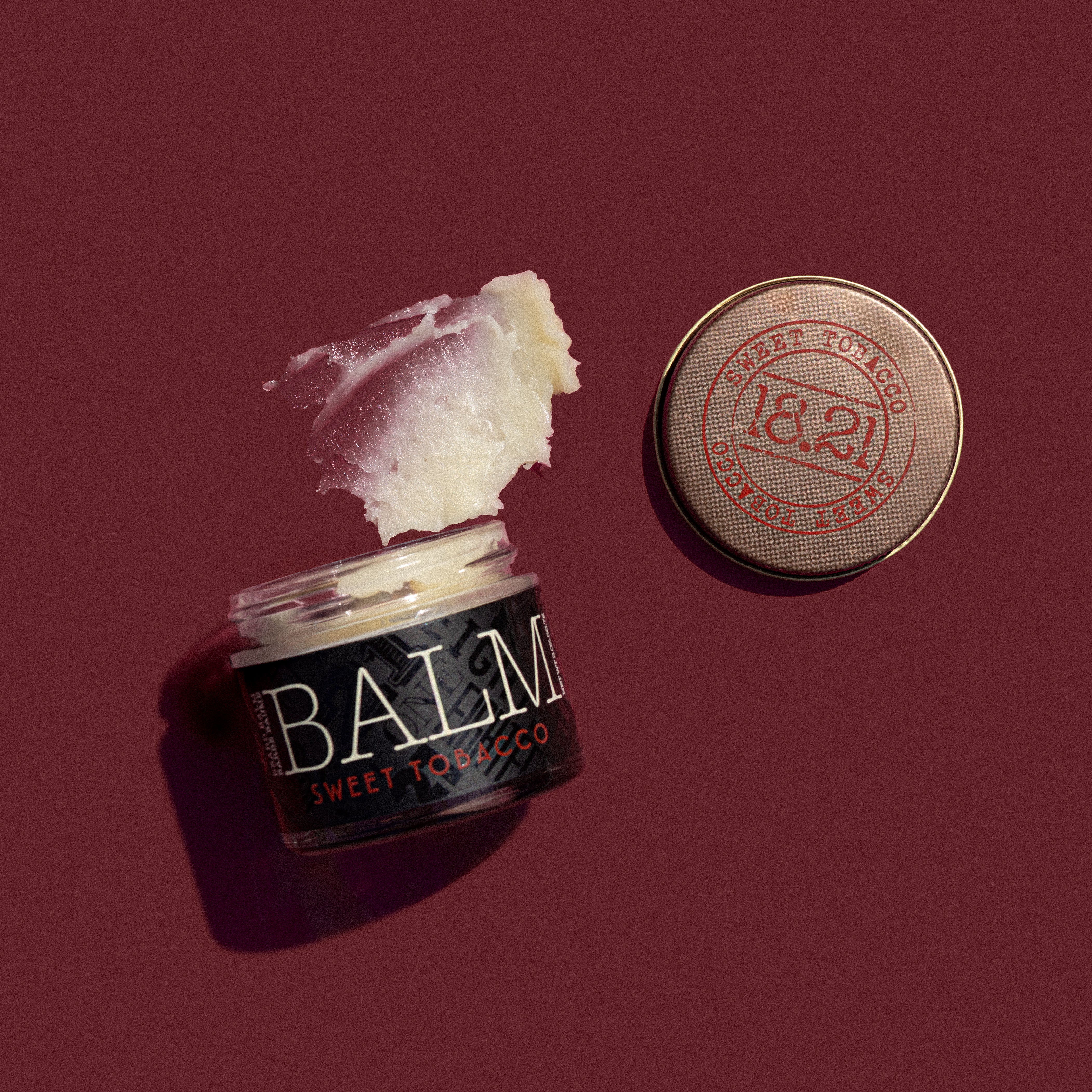 Jar of balm labeled 'Sweet Tobacco' with a red cap on a maroon background