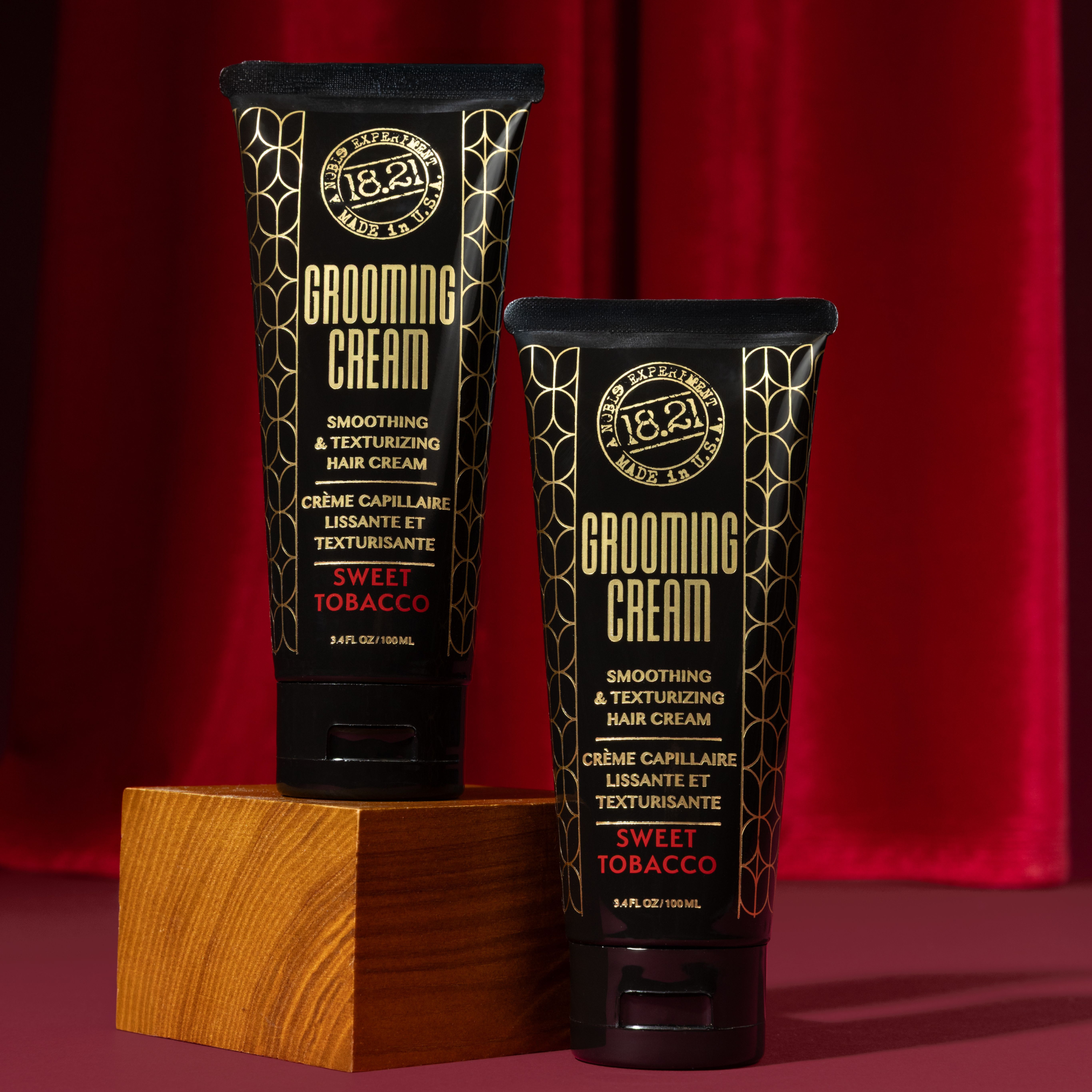 Two tubes of 18.21 Grooming Cream on a wooden block with a red curtain background.