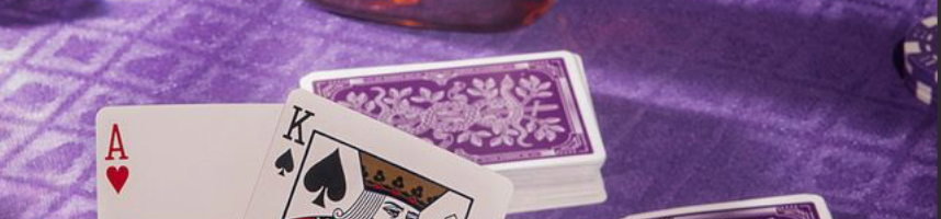 A hand holds overlapping playing cards in the foreground on a purple patterned tablecloth, with a bottle of whiskey, poker chips, and a glass in a dimly lit setting.