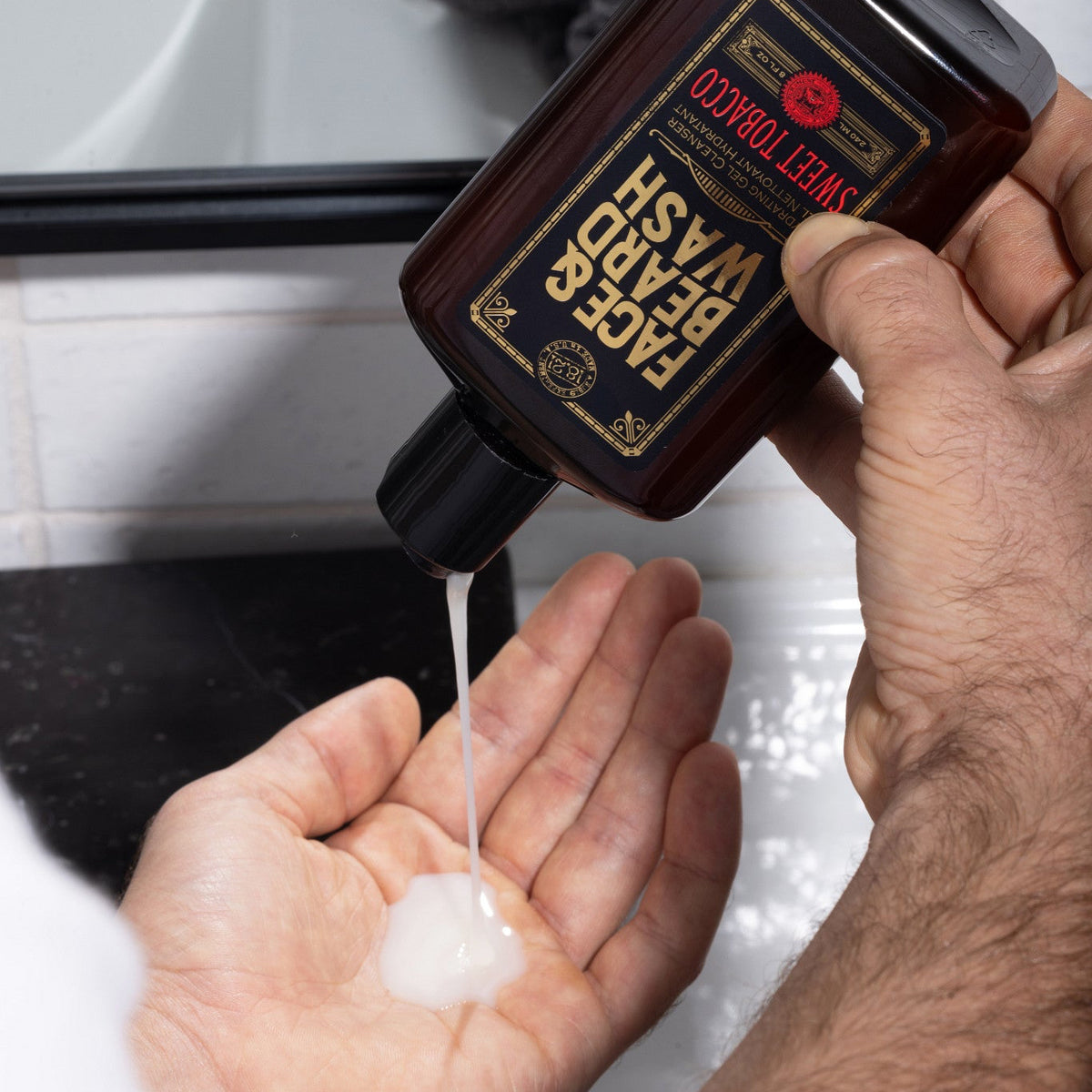 A person holds a dark brown Face and Beard Wash in Sweet Tobacco in the palm of their with the white gel being squeezed onto their hand in a bathroom setting.