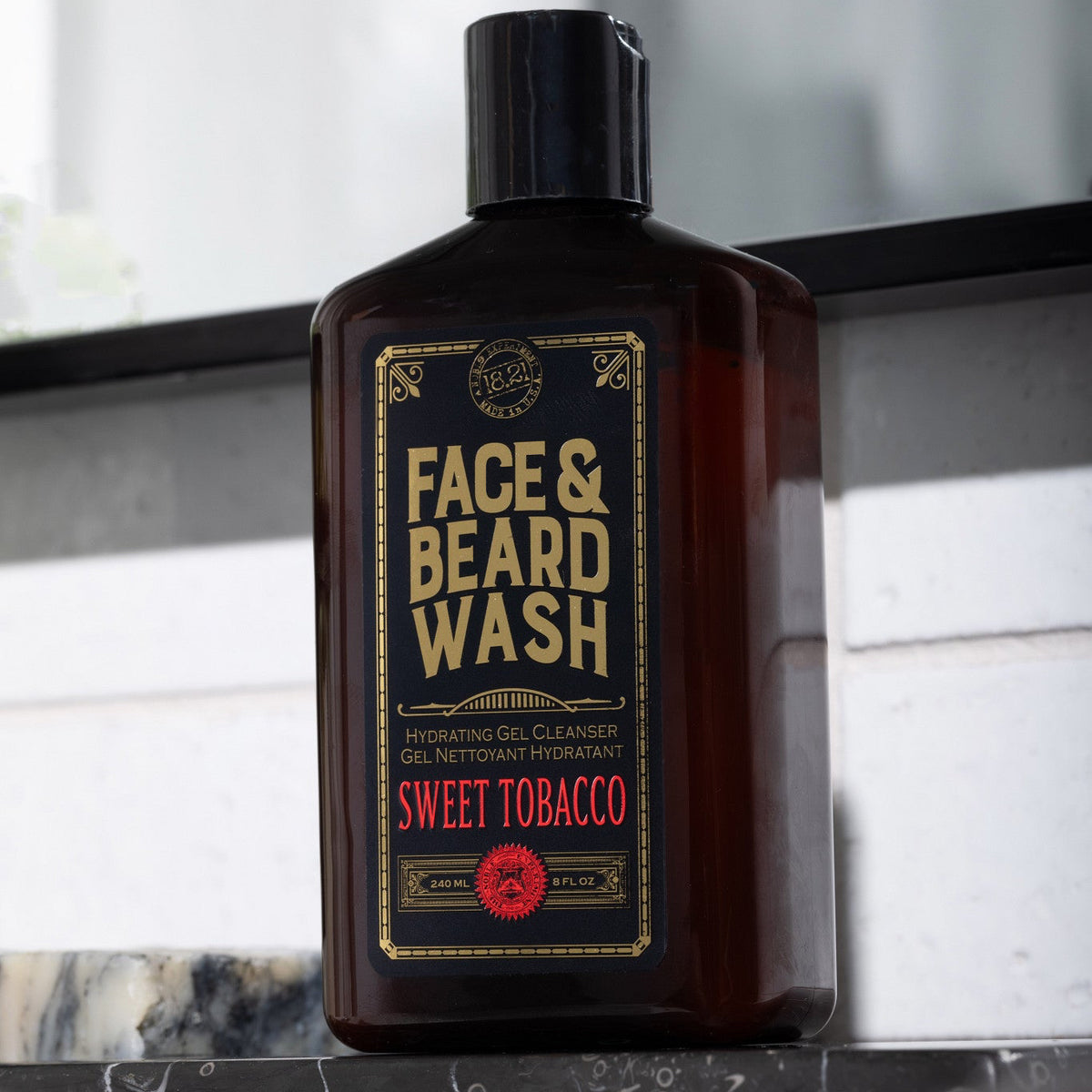 A dark amber bottle of Face & Beard Wash labeled 'Sweet Tobacco' sits on a dark marble surface in front of a white paneled wall.