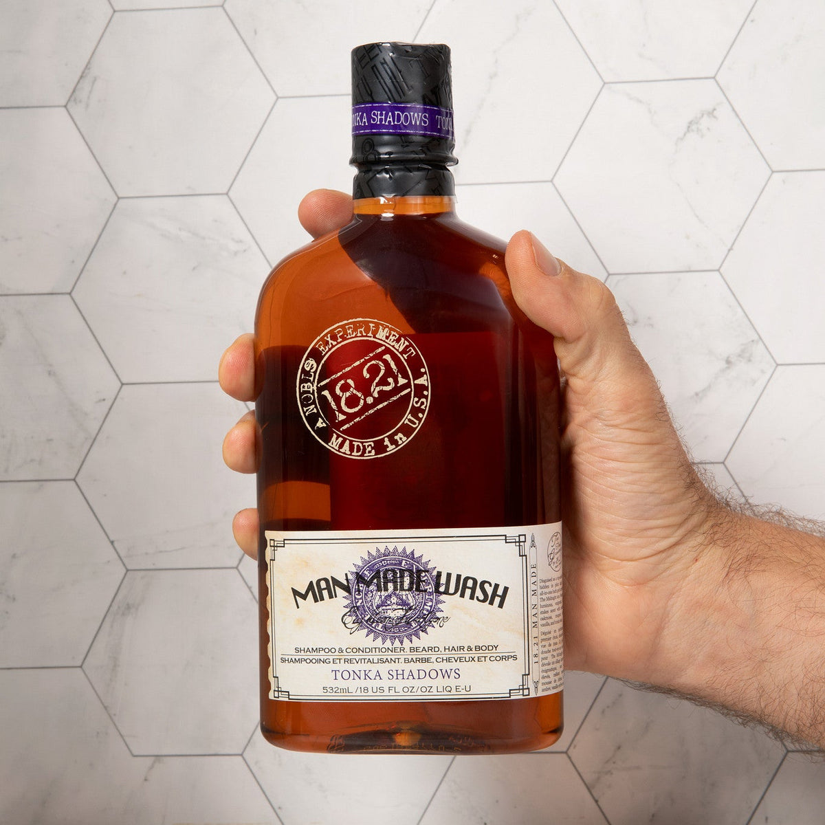 A hand holding a bottle of Men Made Wash Tonka Shadows rum against a white hexagonal tile background.