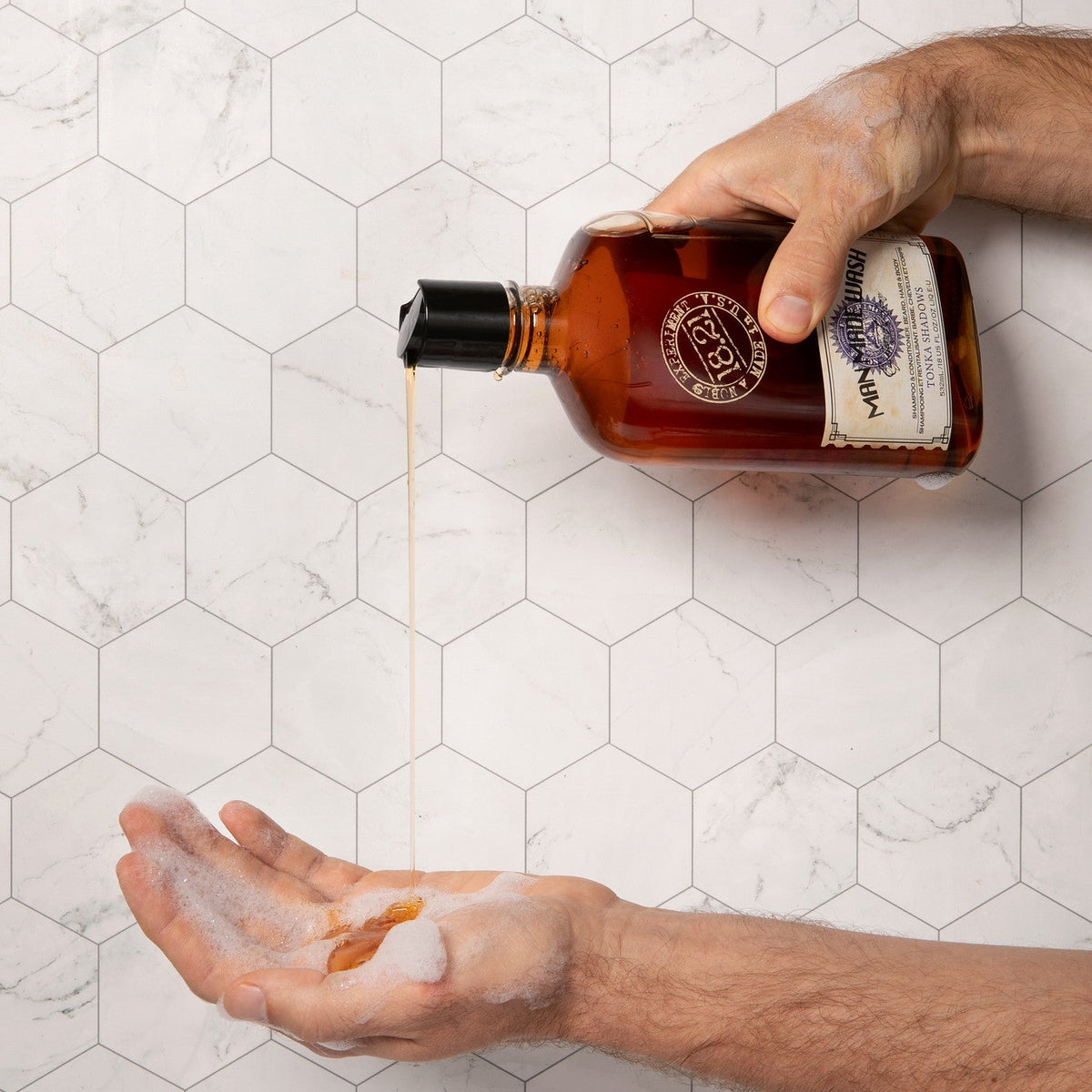 A person's hands holding and pouring an amber bottle of body wash with the oil dripping into the palm of another hand, set against a white hexagonal tile background.