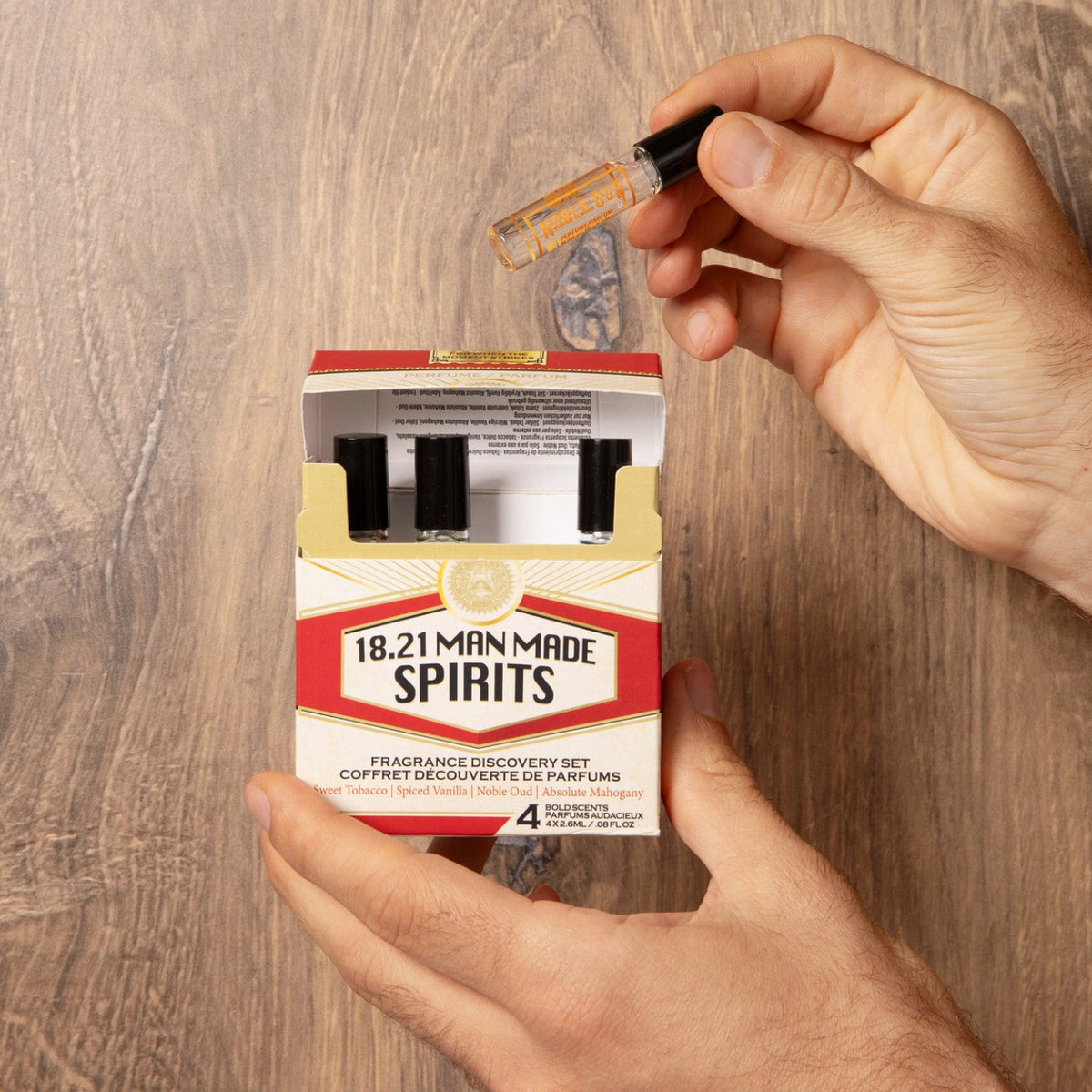 A person's hands holding a 4-pack of 18.21 Man Made Spirits fragrance concentration kits over a wood grain surface, with one hand holding a small glass vial above the open pack.