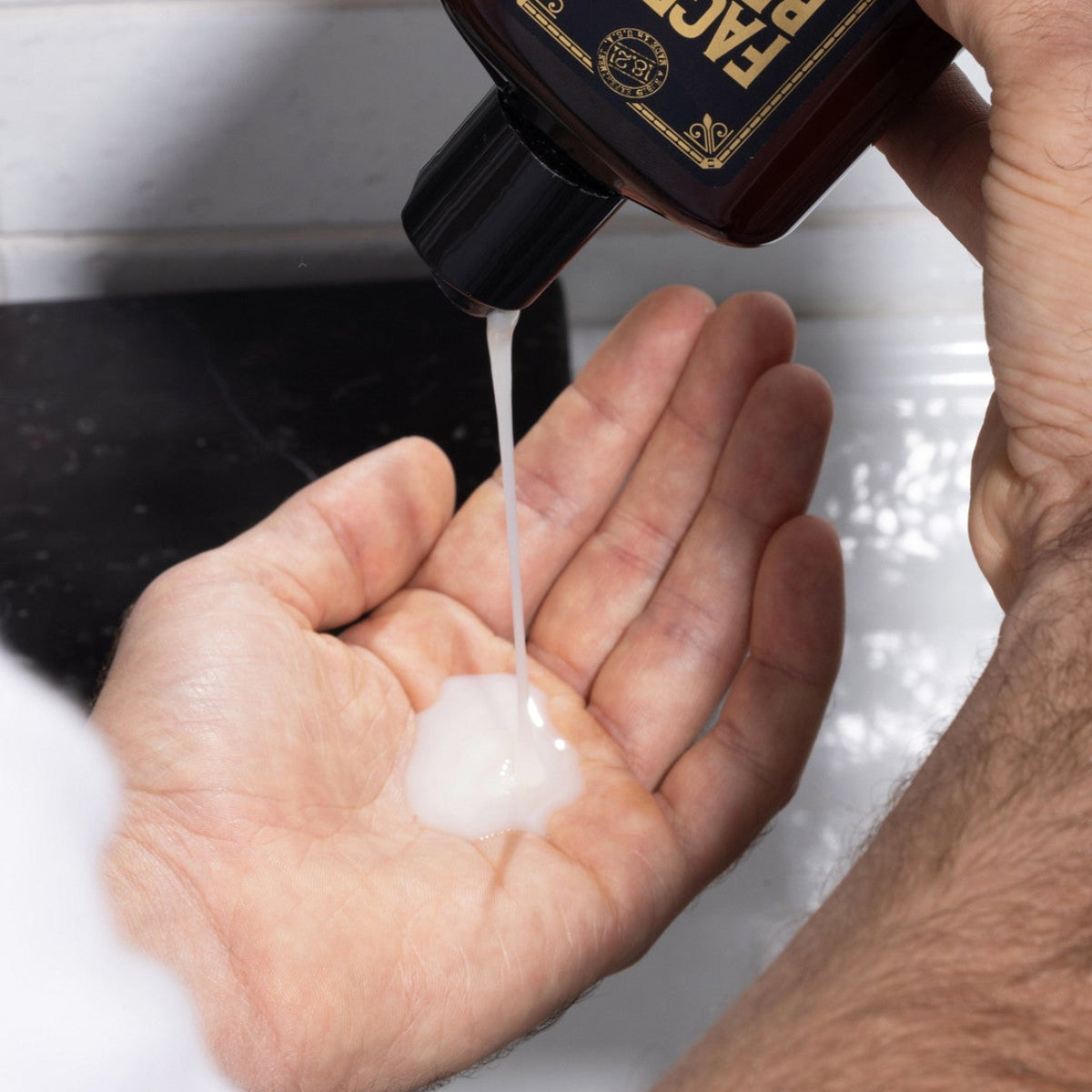 A person holds a dark brown Sweet Escape beard wash bottle over their palm, with the white pearlescent gel cleanser being squeezed onto their hand in a bathroom setting.