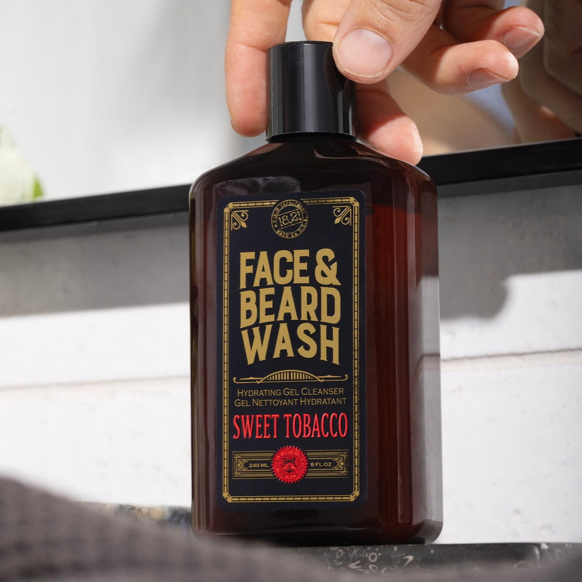 A hand holding a dark amber bottle of Face & Beard Wash, Sweet Tobacco scent, against a blurred bathroom background.