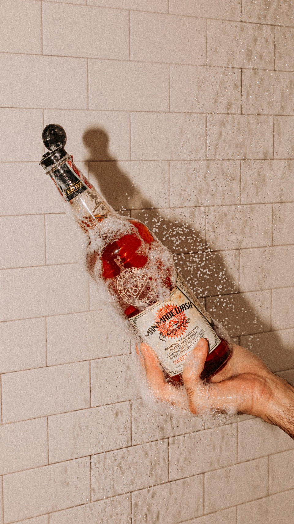 Hand holding 32oz Sweet Tobacco Man Made 3-in-1 Wash in his hand in shower with water 