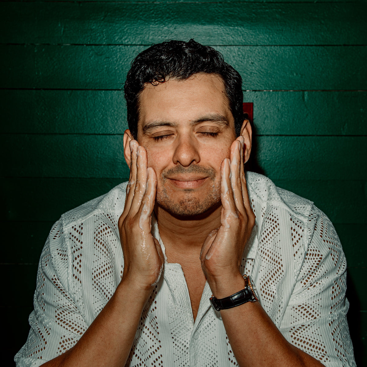A man with closed eyes and hands on his cheeks, wearing a white textured short-sleeve shirt, set against a dark green wooden plank background.
