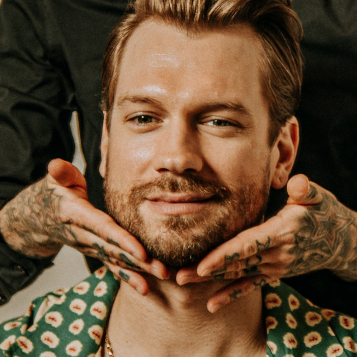 A close-up portrait of a man with styled brown hair and a beard, wearing a green patterned shirt, with tattooed hands holding his jaw. A person in a black shirt is visible behind him.